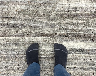 May include: A textured rug in neutral tones, featuring horizontal stripes in grey, beige, and white. The rug's surface appears to be made of woven fibres. The image also shows blue jeans and black socks.