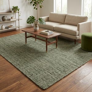 May include: A rectangular area rug with a textured, sage green woven surface. The rug features a dense, looped pile construction and sits flat on a wooden floor, covering a large portion of the space beneath a wooden coffee table and sofa.