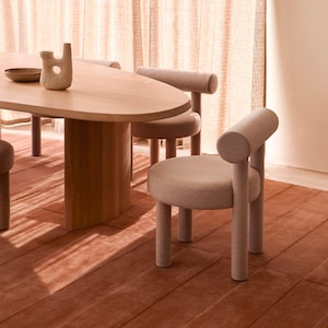 May include: A modern dining room set featuring a light wood oval table and matching chairs. The chairs have rounded backs and legs, upholstered in a neutral fabric. A terracotta-colored rug and beige curtains complete the warm, inviting space.