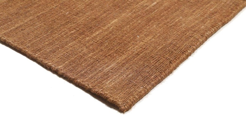 May include: Close-up of a brown woven rug with a textured surface. The rug has a visible weave pattern and a darker brown border. The corner of the rug is visible against a white background.
