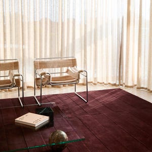 Puede incluir: Escena de salón moderno con una alfombra burdeos oscuro con un patrón geométrico. Se ven dos sillas con marcos metálicos y tapicería beige. Una mesa de centro de cristal con un libro y una esfera decorativa. Cortinas translúcidas.