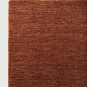 May include: Close-up of a section of a rectangular, rust-colored rug. The rug has a textured, woven appearance with visible horizontal lines. The color is a warm, earthy tone, with a slight variation in shade across the surface. The rug's edge is visible on the left side.