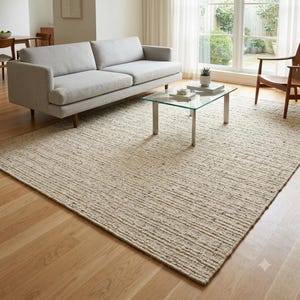May include: A large rectangular area rug featuring a textured, light beige and cream woven pattern with subtle dark flecks throughout. The rug lies flat on a light-toned wooden floor, extending beneath a gray sofa and a glass-topped coffee table.