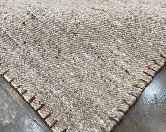 May include: A thick, woven area rug in a neutral beige and cream color palette with a textured, flecked appearance. The edges of the rug are finished with a prominent, dark brown whipstitch detail that runs along the perimeter of the fabric.