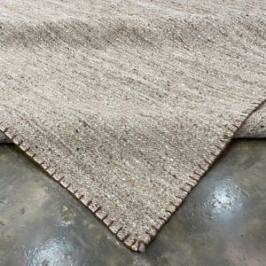 Puede incluir: Primer plano de una alfombra tejida beige con un borde cosido marrón. La alfombra tiene una superficie texturizada con un patrón diagonal. Se ve la esquina de la alfombra, mostrando las costuras detalladas.