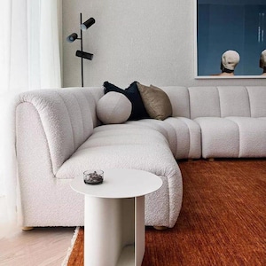 May include: A modern living room with a white sectional sofa, a white side table, and a rust-colored rug. Decorative pillows in navy, olive, and white accent the sofa. A black floor lamp and artwork complete the space.