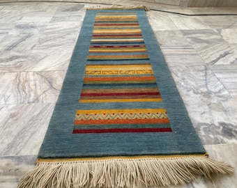 Op de afbeelding: Een blauw rechthoekig tapijt met een centraal ontwerp van horizontale strepen in verschillende kleuren, waaronder geel, oranje, rood en bruin. Het tapijt heeft franjes aan beide uiteinden en ligt op een marmeren oppervlak.