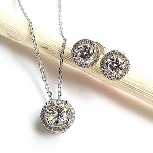 May include: A silver necklace and matching stud earrings set. The necklace has a round pendant with a large, clear gemstone surrounded by smaller stones. The earrings have a similar design. The jewellery is displayed on a white background.