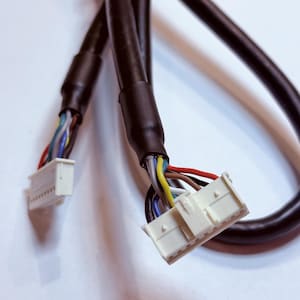 Könnte beinhalten: Nahaufnahme von schwarzen Elektrokabeln mit mehreren farbigen Drähten und weißen Steckern. Die Drähte sind gebündelt und mit den weißen Steckern verbunden. Der Hintergrund ist eine schlichte weiße Oberfläche.