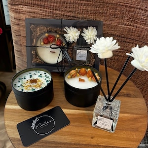 May include: A collection of candles and a diffuser set on a wooden surface. The candles are in black tins, some decorated with dried flowers and crystals. A business card with a logo is also present. The diffuser has white flower accents.