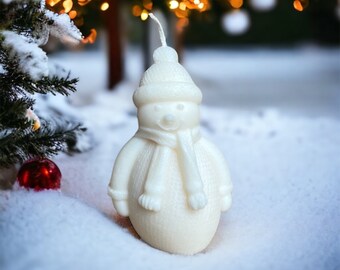 May include: A white snowman candle with a knitted hat and scarf stands in the snow. The candle has a textured surface and a wick at the top. A red ornament and a Christmas tree are in the background.