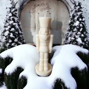 Op de afbeelding: Een crèmekleurige kaars in de vorm van een notenkrakersoldaat staat voor een besneeuwde achtergrond. De kaars heeft een gedetailleerd uniform en hoed. De achtergrond omvat met sneeuw bedekte groenblijvende bomen en een stenen boog.