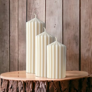 Peut inclure: Trois bougies blanches en forme de pilier avec un motif côtelé, disposées en ordre décroissant de taille sur une souche d'arbre.