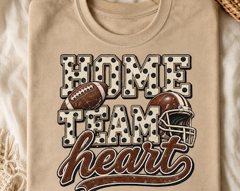 May include: A tan crew neck t-shirt featuring a graphic with the text HOME TEAM in a black and white polka dot block font. A brown football replaces the O in HOME, and a brown football helmet sits next to TEAM. The word heart is in brown script.