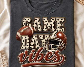 May include: A charcoal gray t-shirt featuring the text GAME DAY in large, white, black-polka-dotted block letters. Below, the word vibes is written in a brown, cursive script. A brown football and a brown football helmet are also displayed.