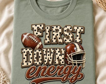 May include: A sage green t-shirt featuring a graphic with the text FIRST DOWN in large cream-colored block letters with black polka dots. A brown football and a brown football helmet are integrated into the design, with the word energy below.