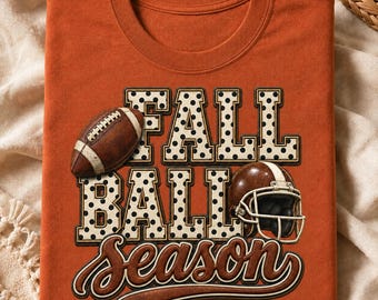 May include: A burnt orange t-shirt featuring the text FALL BALL SEASON in a bold, white, black-dotted font. The design includes a brown football and a brown football helmet with a white stripe, positioned over the lettering on the front.