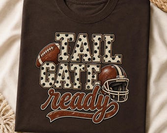 May include: A dark brown short-sleeved t-shirt featuring a graphic print with the text TAIL GATE ready. The design includes a brown football and a brown football helmet. The letters TAIL GATE are filled with a white and black polka dot pattern.