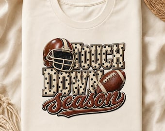 May include: A cream-colored t-shirt featuring a graphic with a brown football helmet, a brown football, and the text "TOUCH DOWN Season" in a blocky, polka-dotted font. The design incorporates brown and cream tones with a vintage-inspired aesthetic.