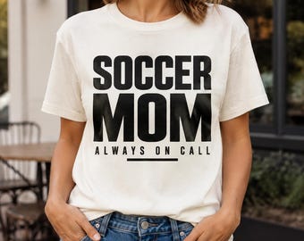 May include: A cream-colored short-sleeve t-shirt featuring the black text SOCCER MOM and the text ALWAYS ON CALL with a horizontal line underneath. The shirt is paired with blue denim jeans with visible metal buttons and rivets at the waist.