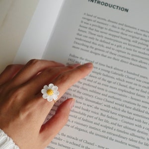 May include: A daisy ring with a white flower and yellow center, set on a gold band. The ring is worn on a finger, with a book open in the background.