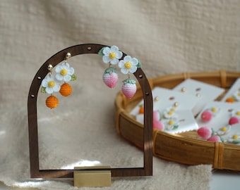 May include: A wooden earring display stand showcasing handmade crochet earrings. The earrings feature white daisy flowers, orange spheres, and pink strawberries. Other earrings are displayed in a basket in the background.