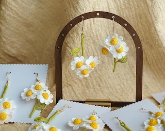May include: Handmade crochet daisy earrings with white petals and yellow centers. The earrings are displayed on white cards and a wooden stand. The stems and leaves are green, and the hooks are gold-colored.