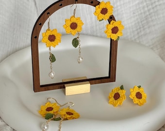 May include: A display of handmade sunflower earrings. The earrings are yellow with a brown center and green leaves. Some earrings have a pearl drop. The earrings are displayed on a wooden stand and a white tray.
