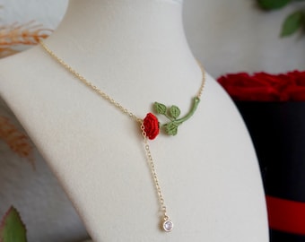 May include: A gold-colored necklace featuring a red crocheted rose and green leaves. A dangling gold chain with a clear gemstone hangs from the rose. The necklace is displayed on a white mannequin.