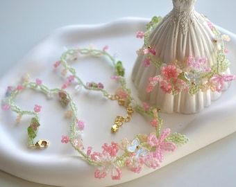 May include: A delicate beaded necklace and bracelet set featuring pink, white, and green floral accents. The jewelry is displayed on a white surface, showcasing the intricate details of the handmade pieces. The necklace has a gold clasp.