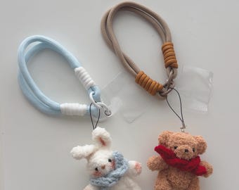 May include: Two handmade hair ties with attached plush charms. One tie is light blue with a white and silver accent, paired with a white bunny wearing a blue scarf. The other is tan with a brown accent, paired with a brown teddy bear wearing a red scarf.