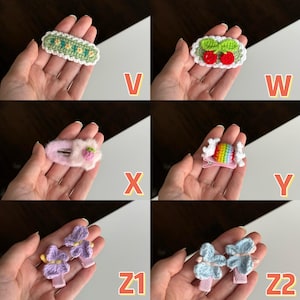 May include: A collection of handmade hair clips in various designs. The clips include a green and white floral design, red cherries, a pink strawberry, a rainbow candy, and purple and blue flower designs. Each clip is held in a hand.