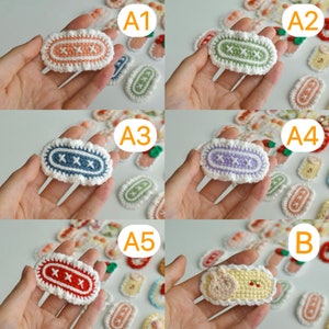 May include: A collection of handmade crochet hair clips in various colors. Each clip features a rectangular design with a white border and an embroidered pattern. Colors include orange, green, blue, purple, red, and yellow. The clips are small and delicate, suitable for various hairstyles.