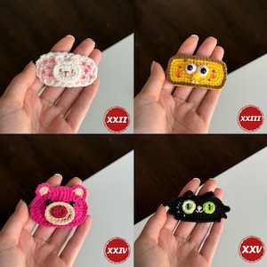 Op de afbeelding: Vier handgemaakte gehaakte haarclips. De clips tonen een wit konijn, een geel wezen met bruine rand, een roze beer en een zwarte kat met groene ogen. Elke clip wordt in een hand gehouden en heeft een rode cirkel met een Romeins cijfer.