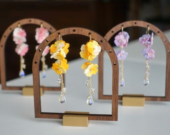 May include: Handmade floral earrings in yellow, pink, and purple displayed on wooden stands. The earrings feature delicate flower designs with gold-colored hooks and dangling crystal accents. The earrings are showcased against a soft background.
