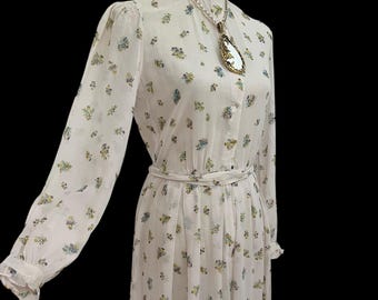 May include: A white floral print dress with long sleeves and a high ruffled collar. The dress features a delicate pattern of blue and yellow flowers and a matching belt. A long pendant necklace with a teardrop-shaped pendant is also visible.
