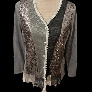 May include: Grey long-sleeved cardigan with a V-neck and button closure. The front features panels of lace, sequins, and black fabric. White lace trim accents the neckline and button placket. The bottom hem has a layered lace design.