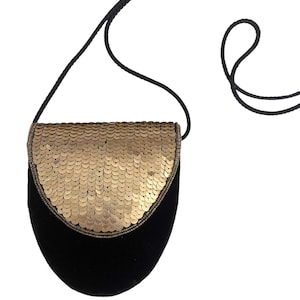 May include: A small black velvet crossbody bag featuring a rounded flap covered in metallic gold sequins. The bag includes a thin, braided black cord strap that extends from the sides, providing a simple and functional design for carrying items.