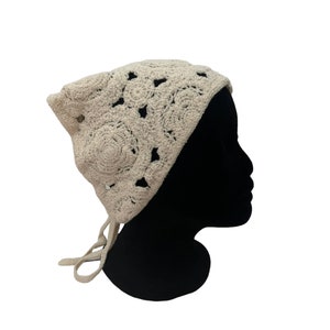 May include: A beige crochet headscarf featuring a repeating floral pattern with openwork details. The headscarf is shaped to cover the top and back of the head and includes two thin crocheted ties extending from the bottom to secure the garment.