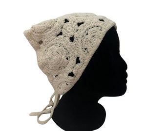 May include: A beige crochet headscarf featuring a repeating floral pattern with openwork details. The headscarf is shaped to cover the top and back of the head and includes two thin crocheted ties extending from the bottom to secure the garment.