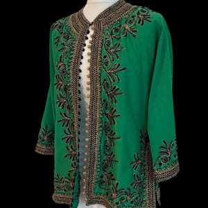 May include: A bright green jacket featuring intricate black and gold embroidery along the front opening, neckline, and cuffs. The garment includes a row of small, round, gold-toned buttons down the centre front and has three-quarter length sleeves.