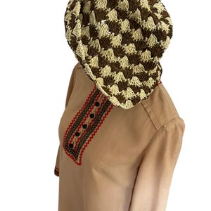 May include: A brown and cream-colored crocheted hat sits atop a tan-colored long-sleeved shirt. The shirt features red and black embroidered detailing around the neckline and sleeves, with black buttons. The hat has a checkered pattern.