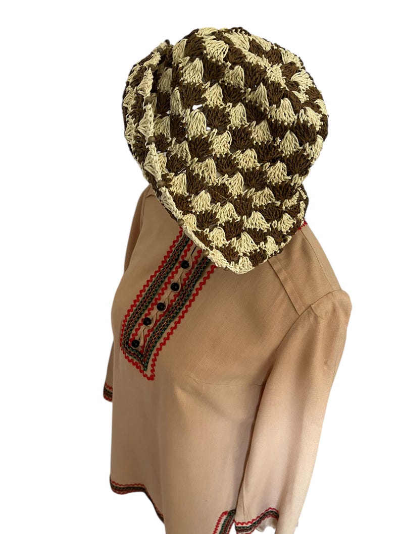 May include: A brown and cream-colored crocheted hat sits atop a tan-colored long-sleeved shirt. The shirt features red and black embroidered detailing around the neckline and sleeves, with black buttons. The hat has a checkered pattern.
