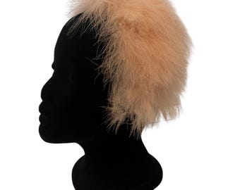 May include: A peach-colored feather headband. The headband is made of soft, fluffy feathers and is worn on a black mannequin head. The feathers are a light, pastel peach color, and the headband is a stylish accessory.