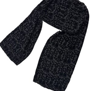 May include: A dark gray scarf with a black floral pattern. The scarf is made of a soft, velvety material and is draped in a curved shape. The floral design is repeated across the entire scarf.