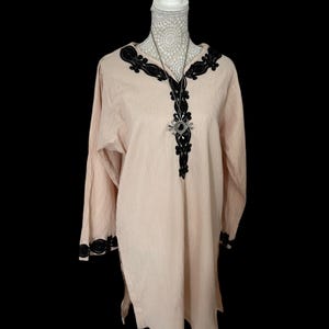 May include: A long-sleeved, light pink tunic with black embroidery around the neckline and cuffs. The tunic has a V-neck and a long, flowing silhouette.