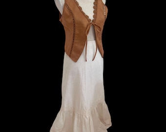 May include: A brown suede vest with a scalloped neckline and tie closure, paired with a cream-colored maxi skirt featuring tiered ruffles. The outfit is displayed on a mannequin against a black background, showcasing a bohemian style.