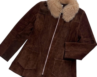 Könnte beinhalten: Eine dunkelbraune Wildlederjacke mit beigem Kunstfellkragen, einem durchgehenden silberfarbenen Reißverschluss und zwei großen aufgesetzten Taschen auf der Vorderseite. Die Jacke hat lange Ärmel und eine strukturierte Passform mit sichtbaren vertikalen Nähten.