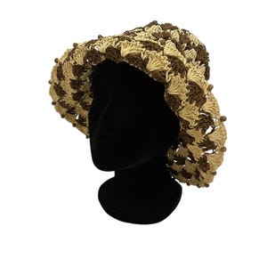 May include: A handmade crochet hat in a brown and beige pattern. The hat features a wide brim and is adorned with small, decorative beads around the edge. The hat is displayed on a black mannequin head against a white background.