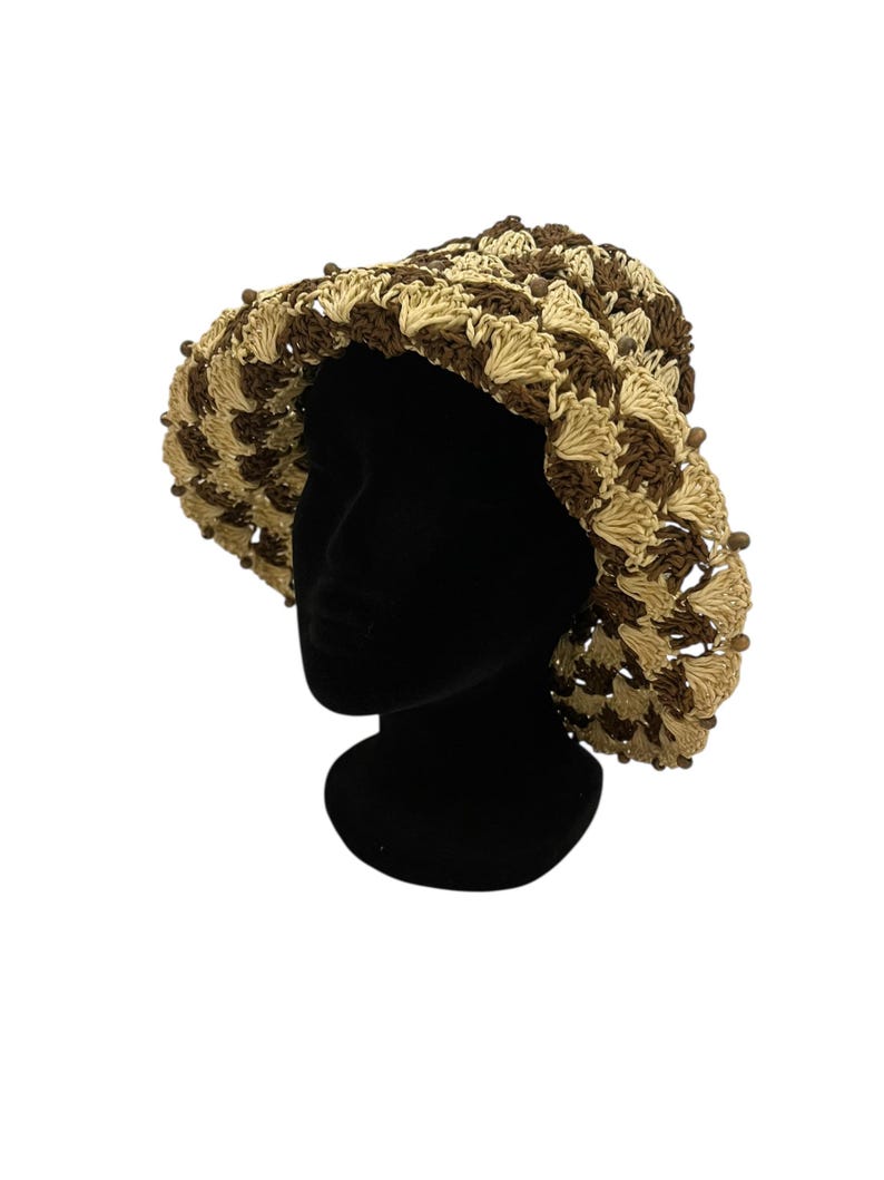May include: A handmade crochet hat in a brown and beige pattern. The hat features a wide brim and is adorned with small, decorative beads around the edge. The hat is displayed on a black mannequin head against a white background.